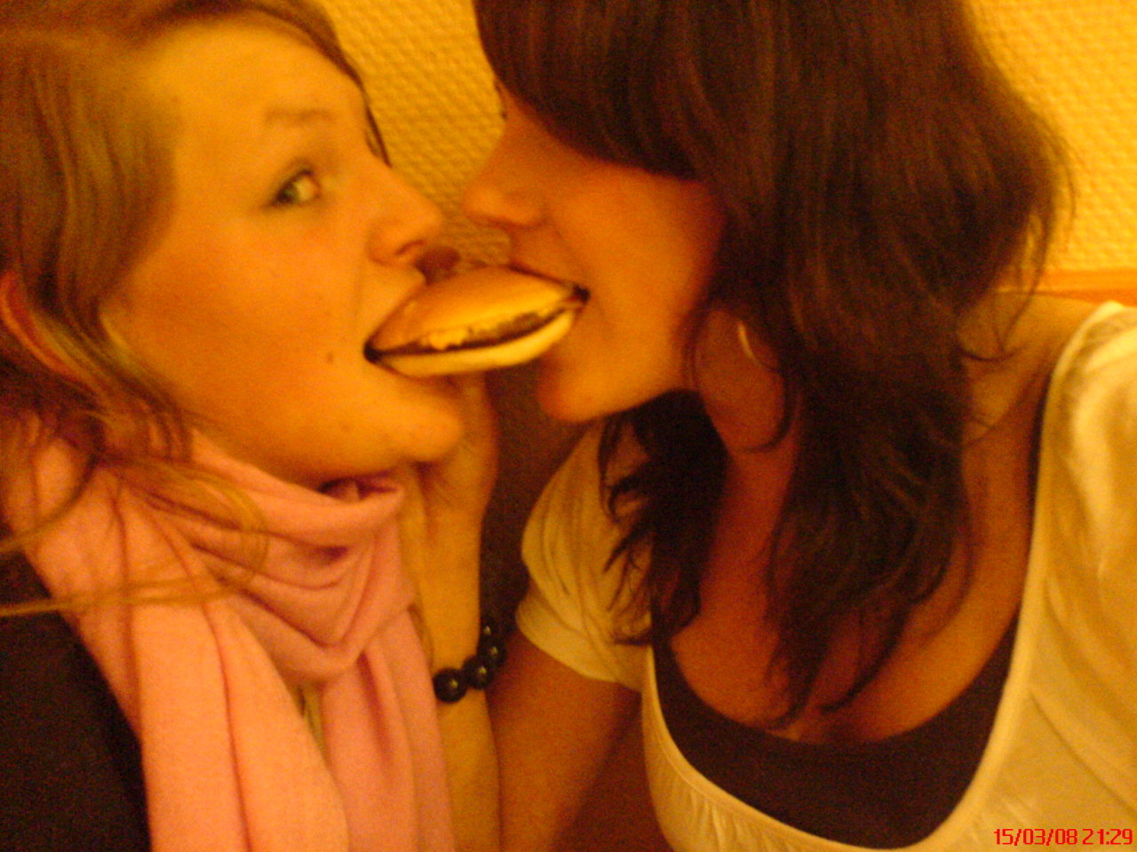 Two girls awkardy take a bite of the same burger, they are laughing. The camera text at the bottom reads: 15/03/08 21:29