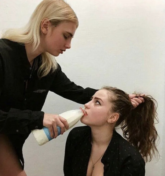 Two girls. One of them seems to be on her knees, the other holds her ponytail while making her chug a bottle of milk. They stare deeply at eachother's eyes.